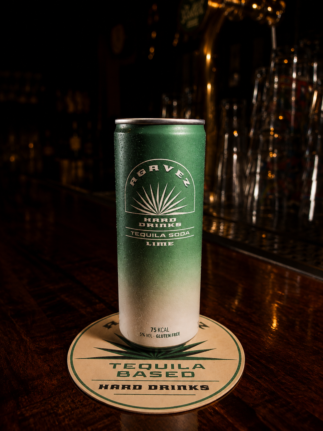 An Agavez Tequila Soda Lime can on a coaster on a dark bar top with warm lights behind